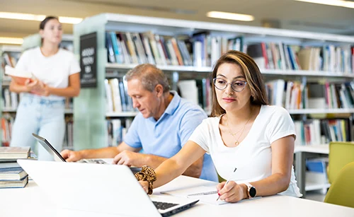 Adult students studying in a library at Amarillo College, with a woman working on a laptop and a man reading at a table during a GED and ESL class.