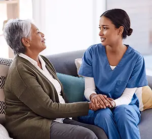 Image shows young female Mental Health Technician holding hands with a senior woman and on sofa