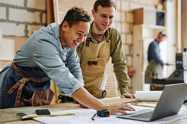 Image shows two individuals collaborating in a workshop environment, looking at plans on a laptop