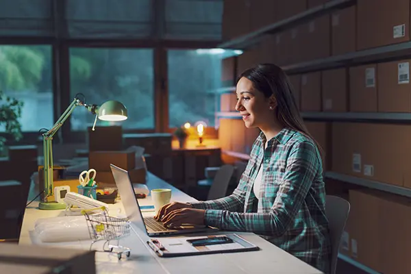Image shows a young woman working on a laptop at a desk in a dimly lit office environment