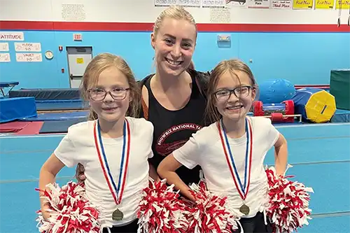 Third slide AC gymnastics instructor standing with two youngs students holding pompoms