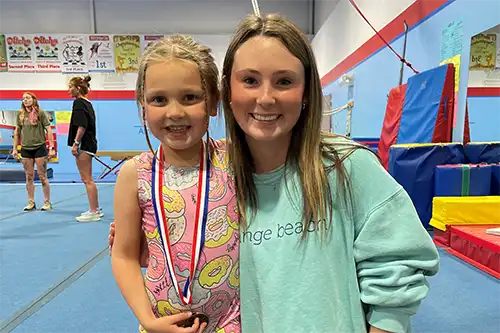 Fourth slide AC gymnastics instructor standing next to a young student wearing a medal